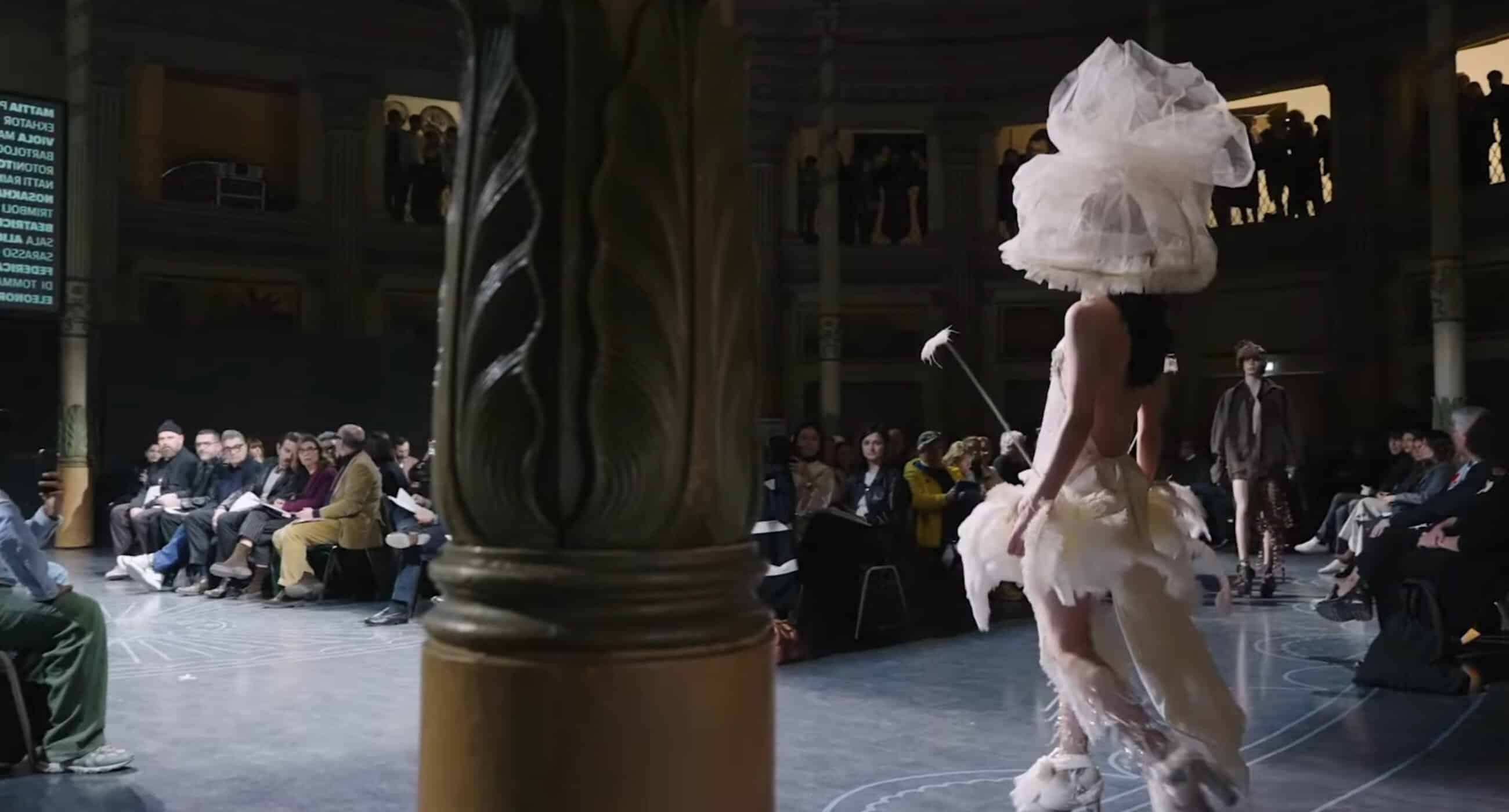 Model in an elaborate white outfit and headpiece walks on a circular runway with seated audience in a domed venue.