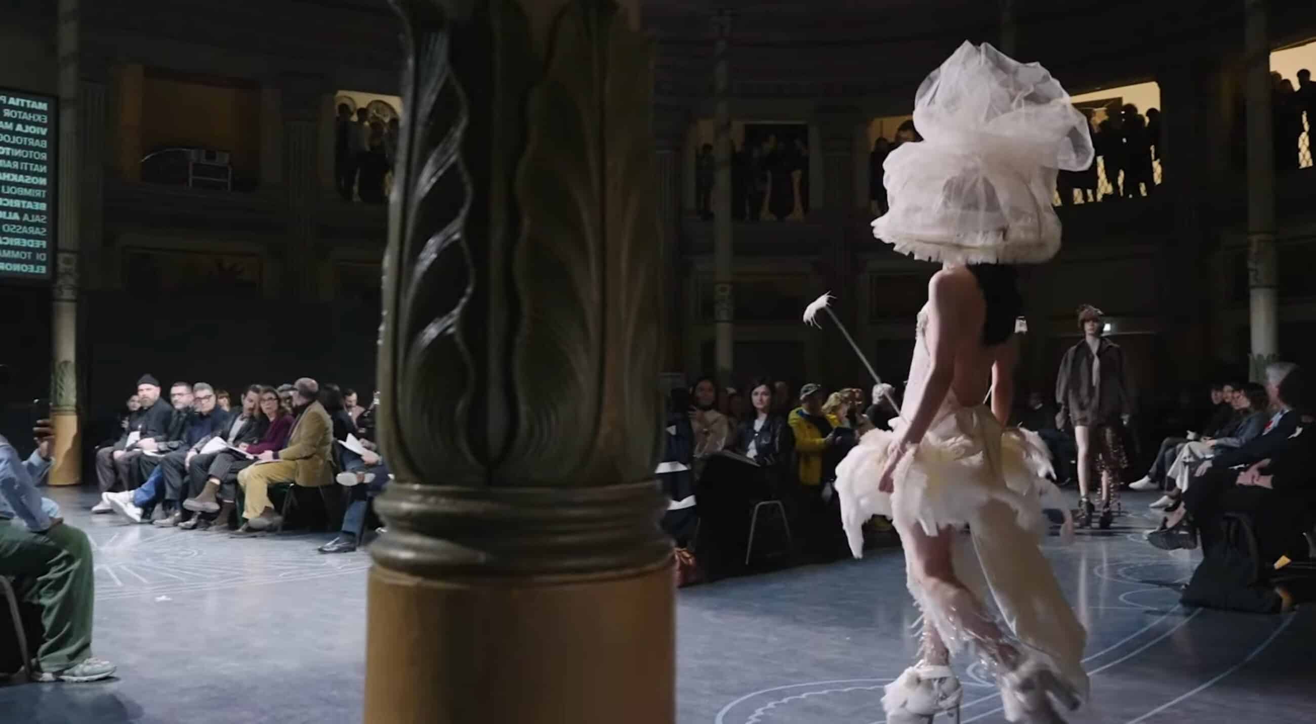 Model in an elaborate white outfit and headpiece walks on a circular runway with seated audience in a domed venue.