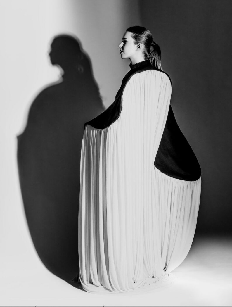 Black and white image of a woman in a long, flowing dress posing dramatically in profile. Her shadow is cast on the wall behind her.