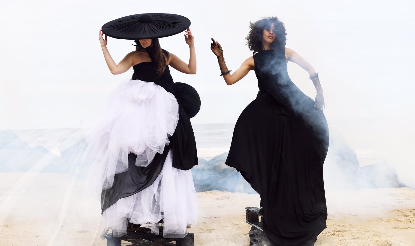 Two women in black and white avant-garde dresses stand on a sandy beach. One wears a large hat, the other has curly hair. There's a misty atmosphere around them.