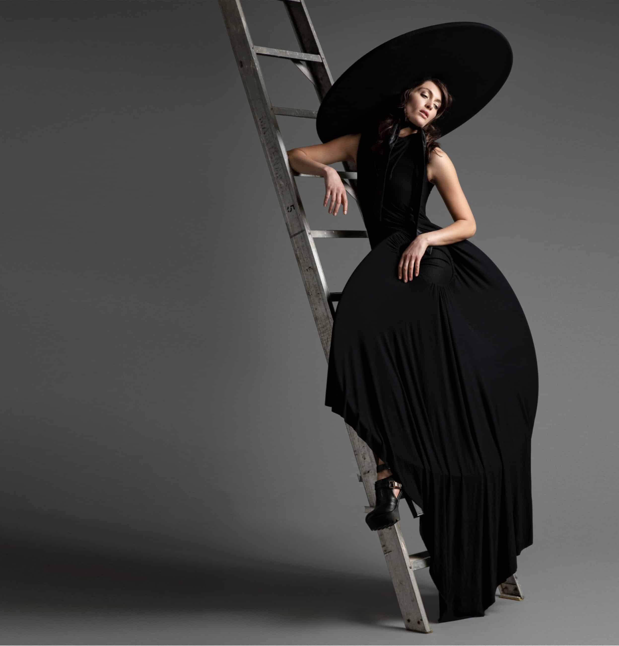A woman in a long black dress and wide-brimmed hat sits on a ladder against a gray background, exuding a dramatic and stylish pose.