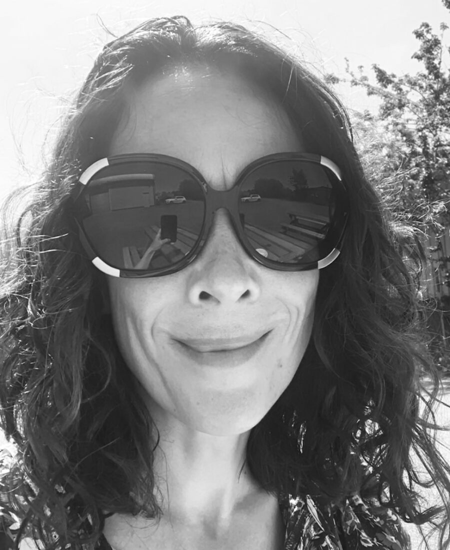 Woman with curly hair wearing large sunglasses, smiling at the camera outdoors. Black and white image.