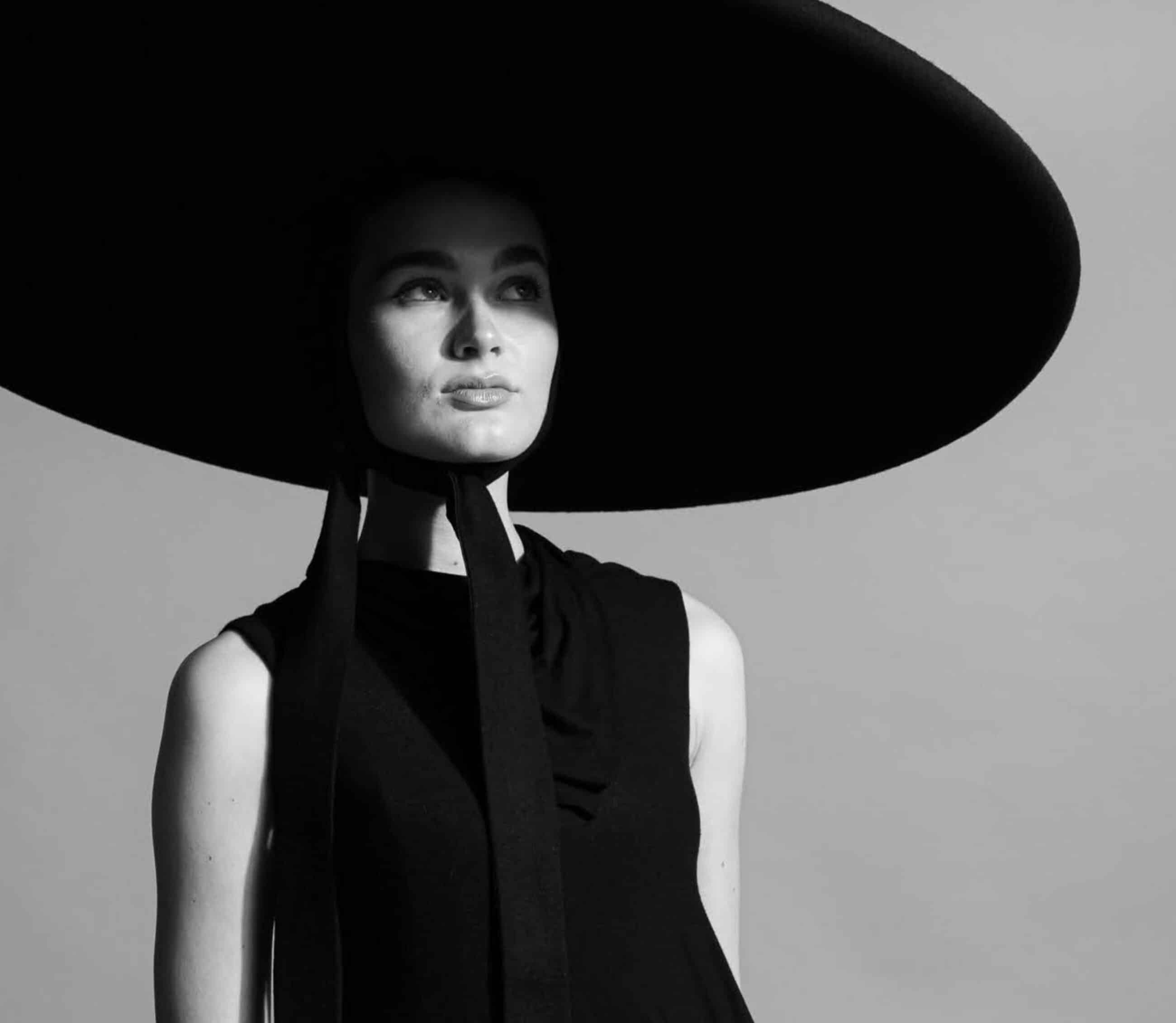 Person in black outfit wearing a large, wide-brimmed hat against a plain background, staring to the side.
