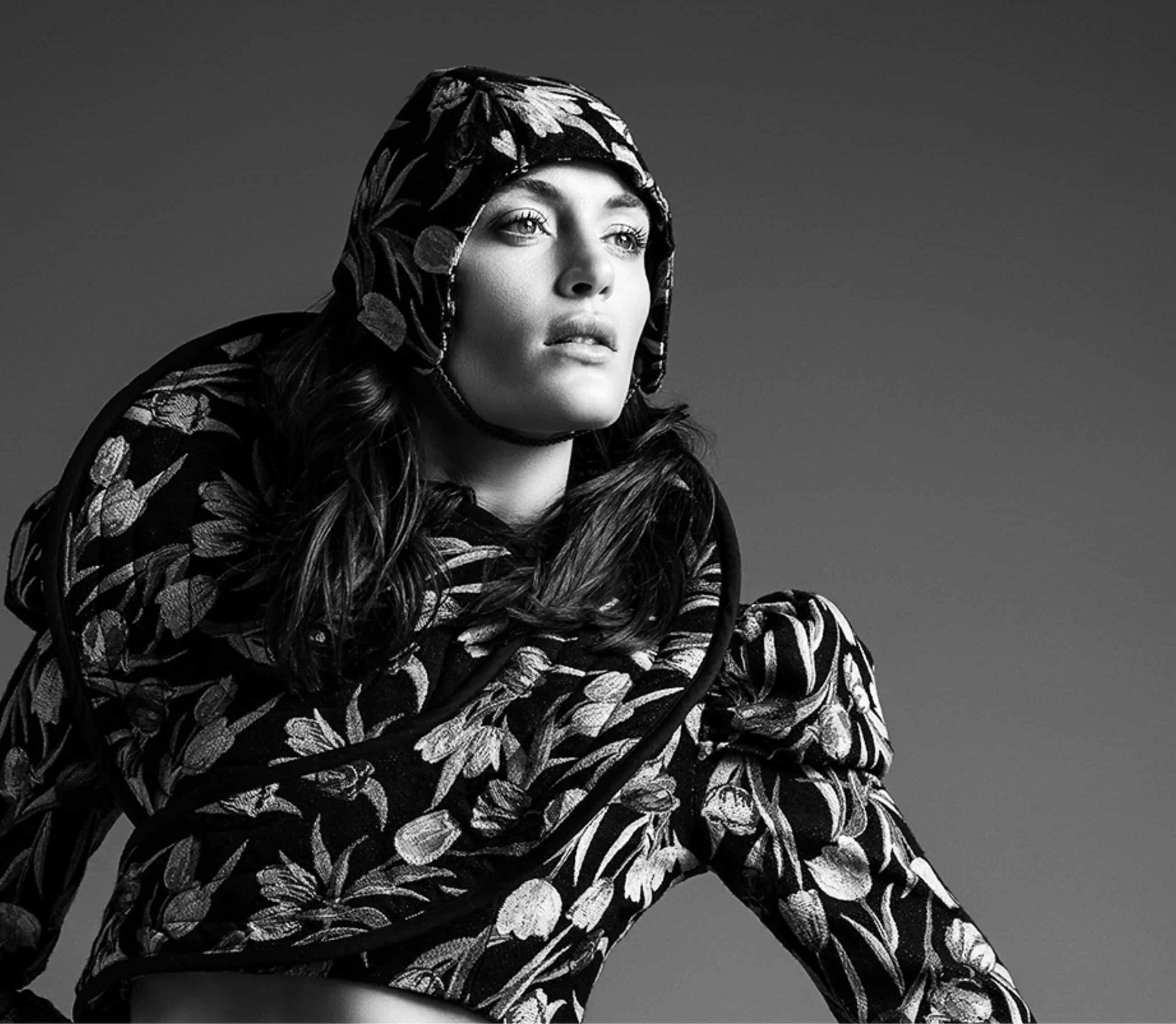 Person wearing a floral-patterned hooded outfit, gazing upwards in a studio-lit setting. Black and white image.