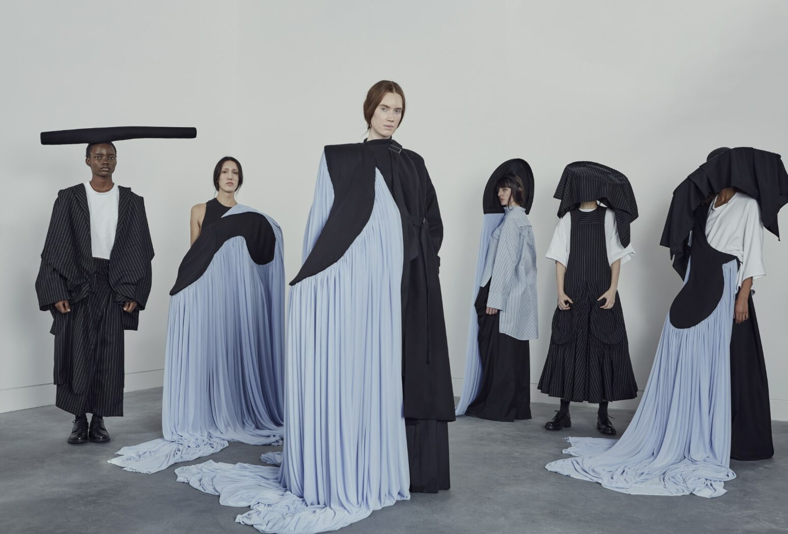 Six people wear avant-garde, flowing black and blue outfits with abstract designs and headpieces, standing in a neutral room.