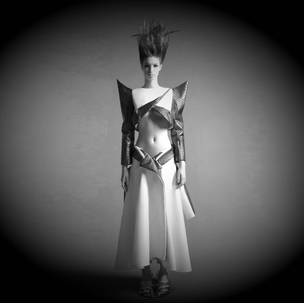 A model in a futuristic outfit with structured, angular shoulders and a high-waisted skirt poses against a simple backdrop. Her hair is styled upwards dramatically. Image is in black and white.
