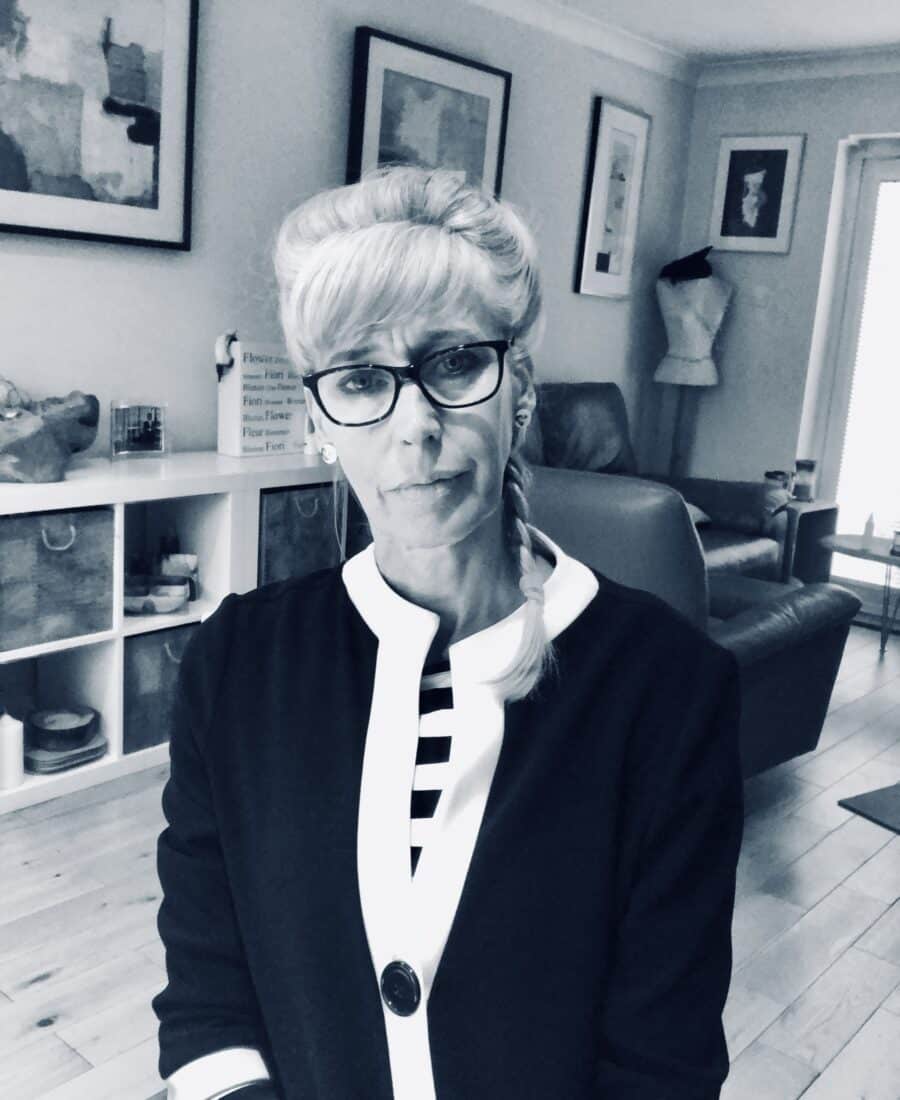 A woman with glasses and a braided hairstyle sits indoors, wearing a black and white striped top. The room has framed artwork on the walls and shelves with various items.