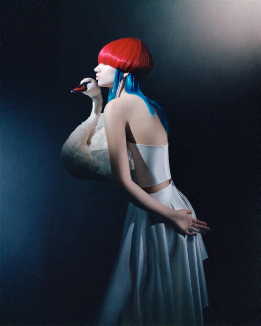 A woman with red and blue hair in a white dress holds a swan against a dark background.