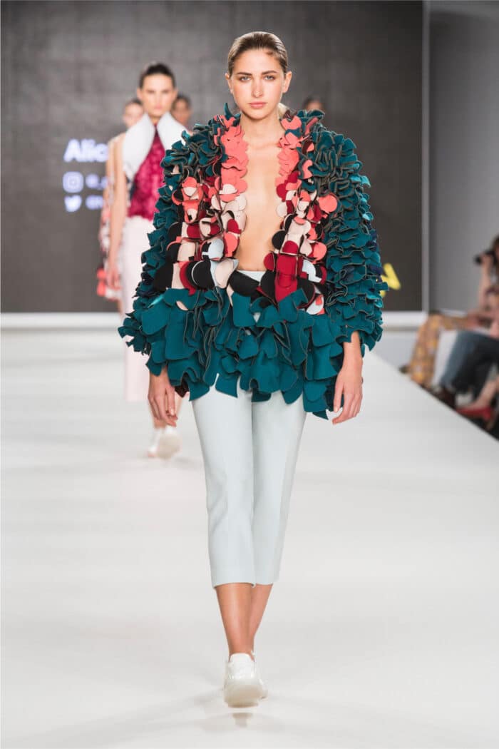 Model walking on runway wearing a bold, multi-colored ruffled top and light blue pants, with audience in background.