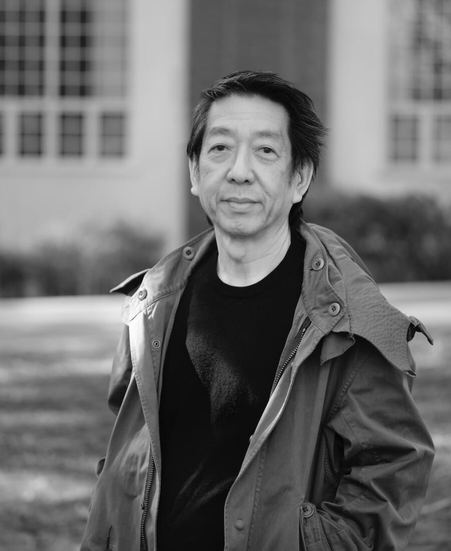 A person with dark hair wearing a jacket stands outdoors in front of a building.