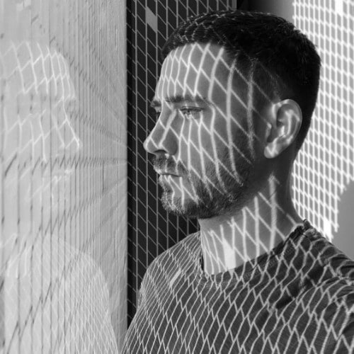 Man in a T-shirt is standing by a window with a grid pattern of shadows cast on his face and body, looking outside.