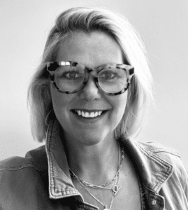 Woman with short blonde hair, wearing large glasses, a denim jacket, and layered necklaces, smiling in a black and white photo.