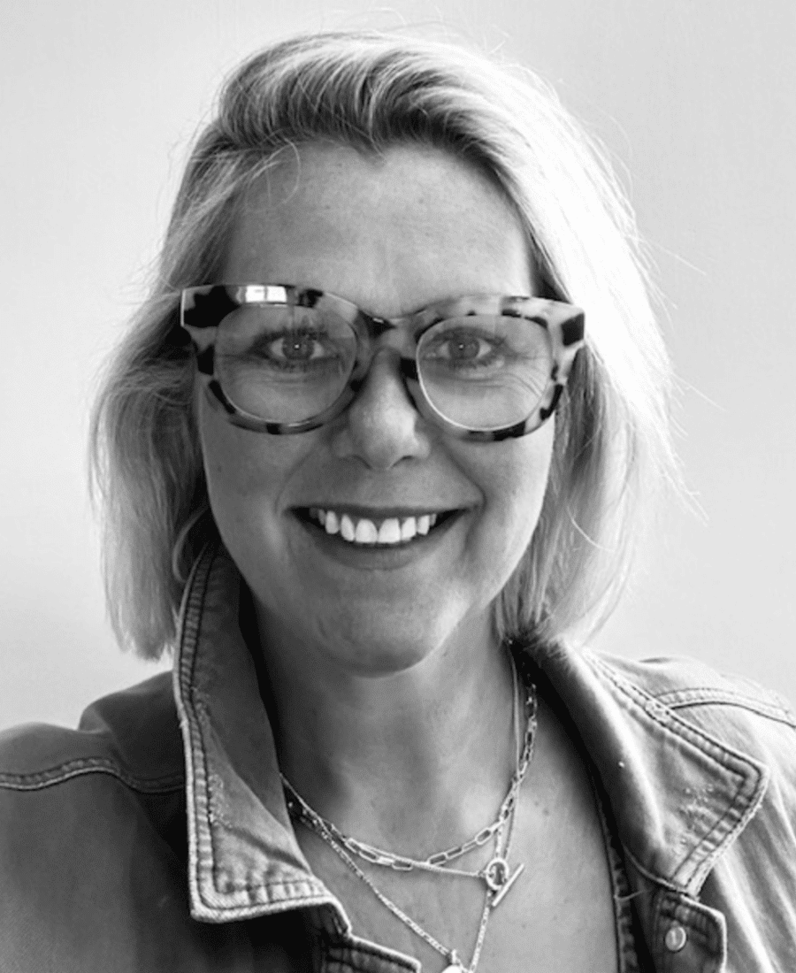 Woman with short blonde hair, wearing large glasses, a denim jacket, and layered necklaces, smiling in a black and white photo.