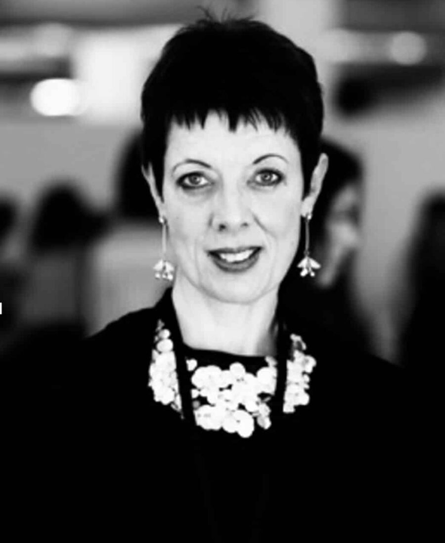 A woman with short dark hair, wearing a floral necklace and earrings, stands in a black and white setting.