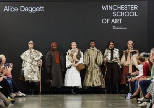 Six models display fashion designs on a runway at Graduate Fashion Week 2024. Designer Alice Daggett's name is displayed on the backdrop. Audience members are seated on both sides of the runway.