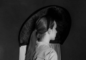 Person in profile wearing a large, circular hat and striped shirt with back detail against a dark background.