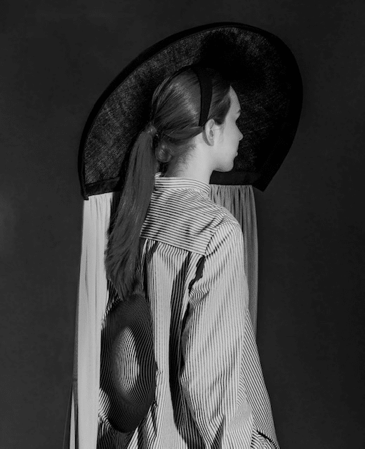 Person in profile wearing a large, circular hat and striped shirt with back detail against a dark background.