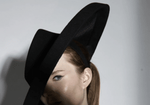 Person wearing a large, black hat with a wide brim and dark clothing, partially shadowed, looking off-camera.