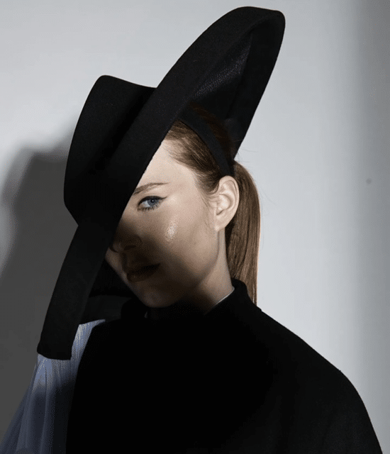 Person wearing a large, black hat with a wide brim and dark clothing, partially shadowed, looking off-camera.