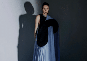 Person in a dramatic floor-length gown with a blue draped skirt and black top. Shadow cast on wall in studio setting.