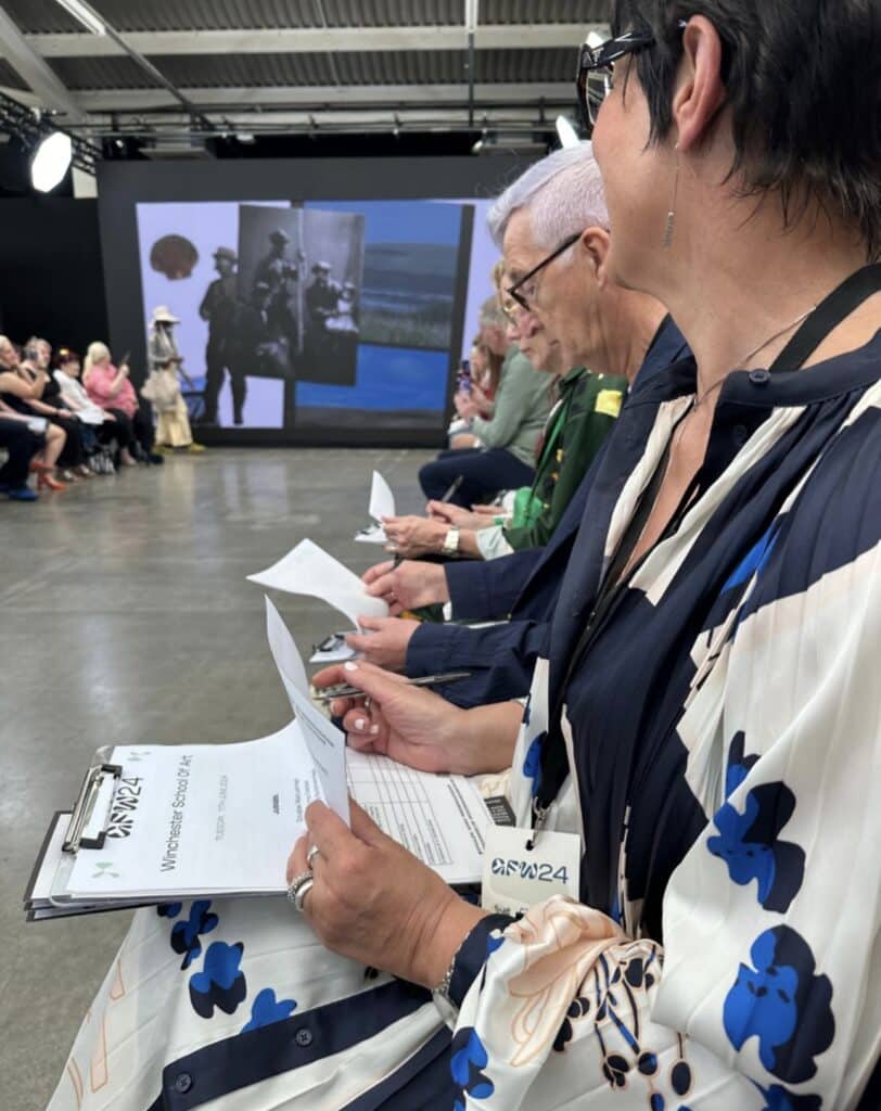 People sit in a row with papers and clipboards, watching a presentation on a large screen in a modern indoor setting.