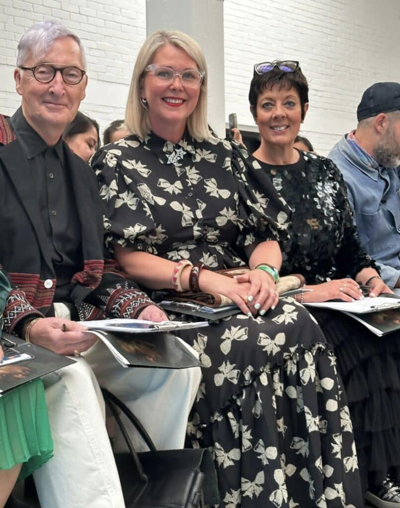 Three people sitting in a row at an indoor event, smiling and holding papers. Two women are in printed dresses, and a man is in glasses and a patterned jacket.