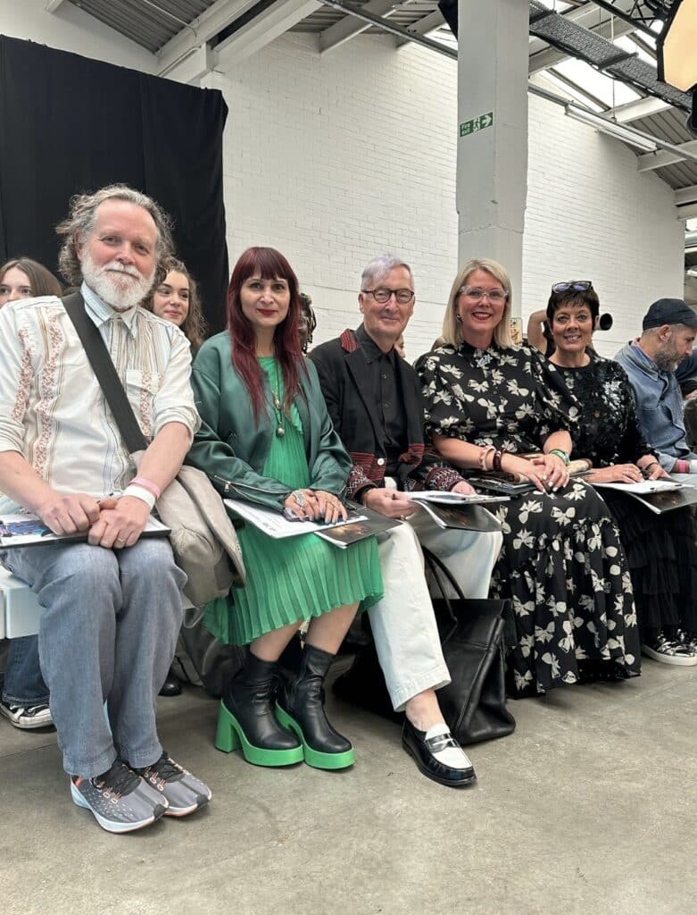 A group of five people sitting on a bench at a fashion show, dressed in various styles, with a brick wall and black curtain in the background.