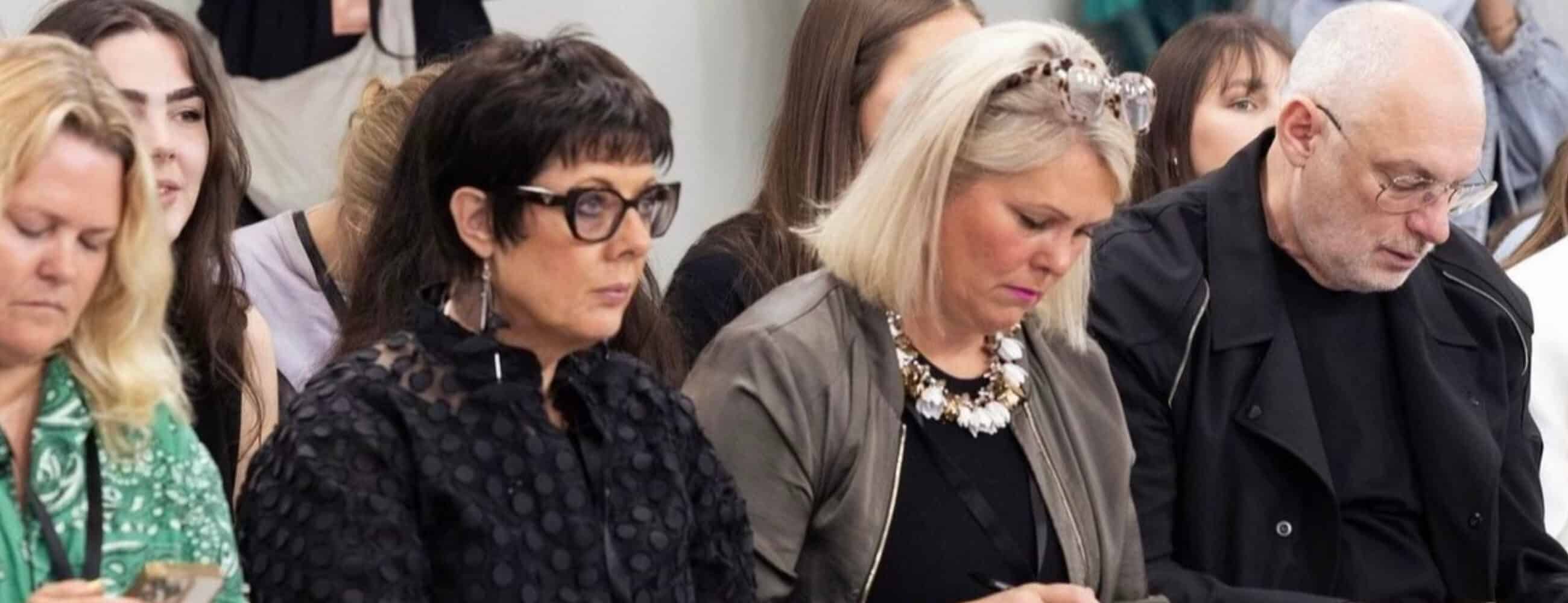 A group of people sitting closely together, some are looking down or at their phones, in what appears to be an indoor event.