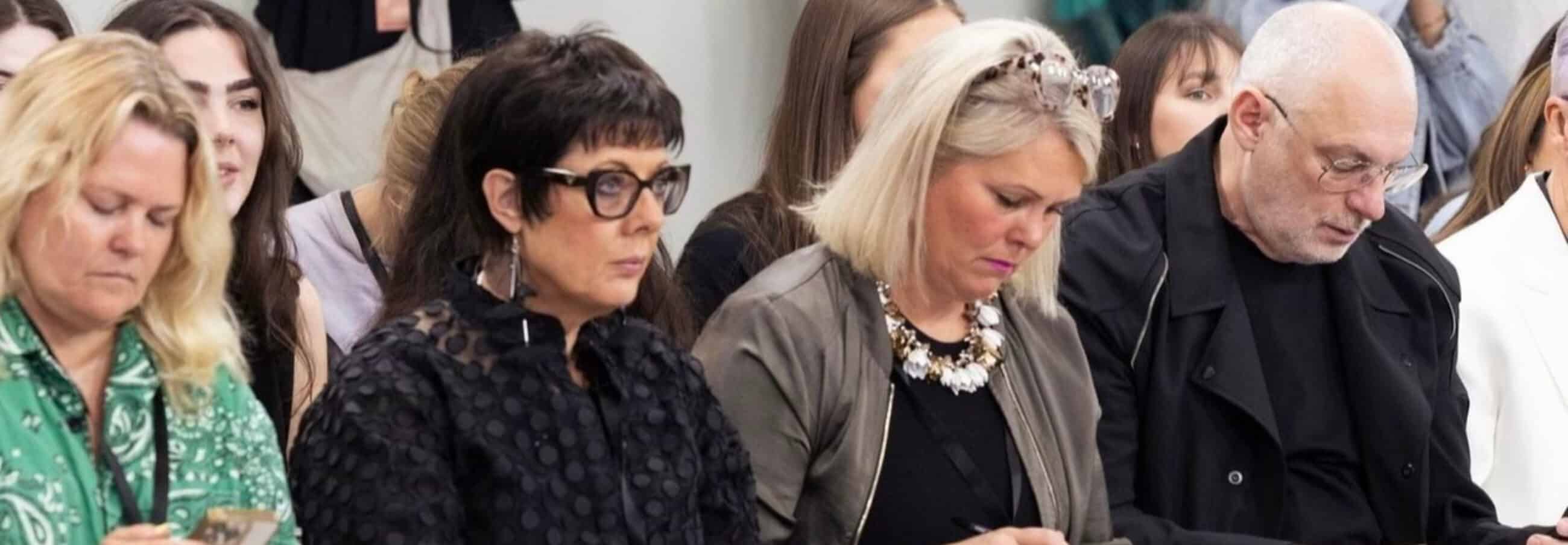 A group of people sitting closely together, some are looking down or at their phones, in what appears to be an indoor event.