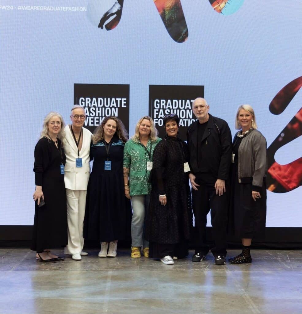 A group of seven people standing in front of a screen displaying "Graduate Fashion Week" signs. They are posing for a photo and appear to be at an event or conference.