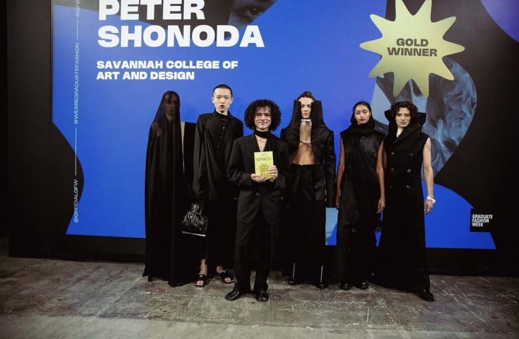 A group of people in formal and avant-garde attire stand in front of a blue and black backdrop featuring the name Peter Shonoda and a "Gold Winner" badge.