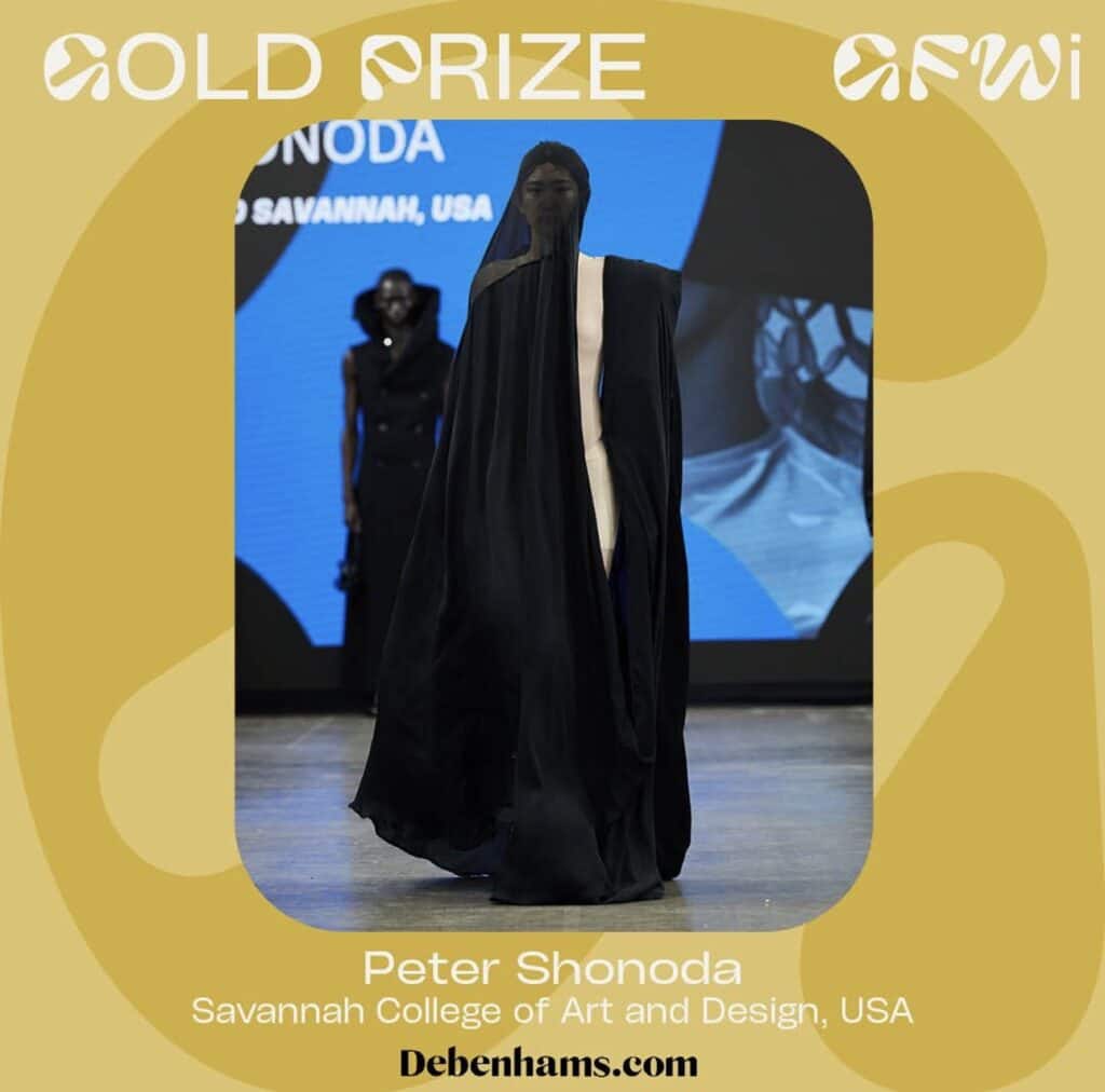 A model walks the runway in an elaborate black outfit featuring a full-face covering at a fashion show, with a gold prize label in the background.
