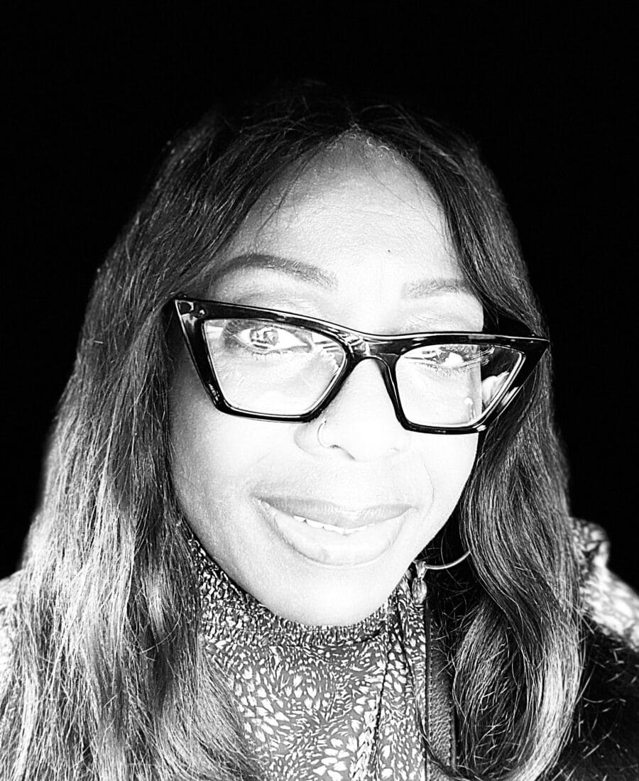 Black and white photo of a person with long hair and glasses, smiling directly at the camera.