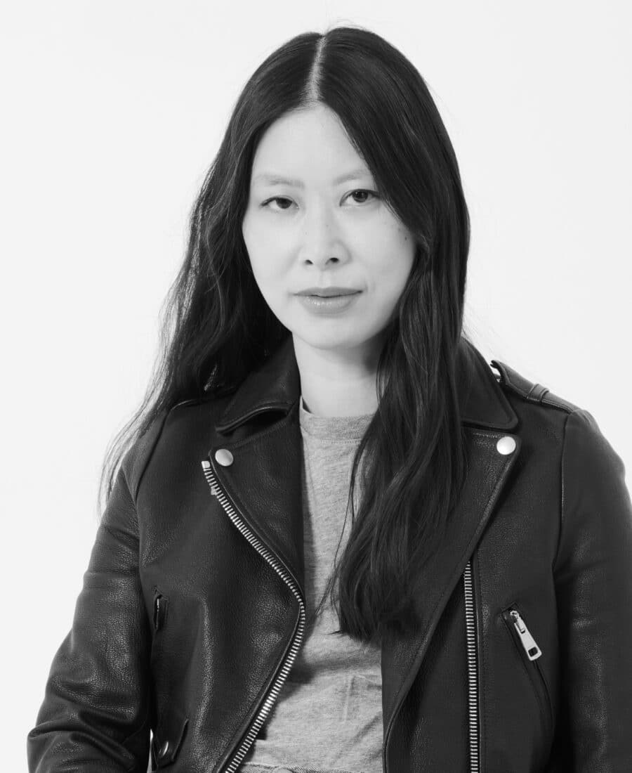 Person with long hair wearing a leather jacket over a gray shirt, facing the camera with a neutral expression, in a black and white photo.