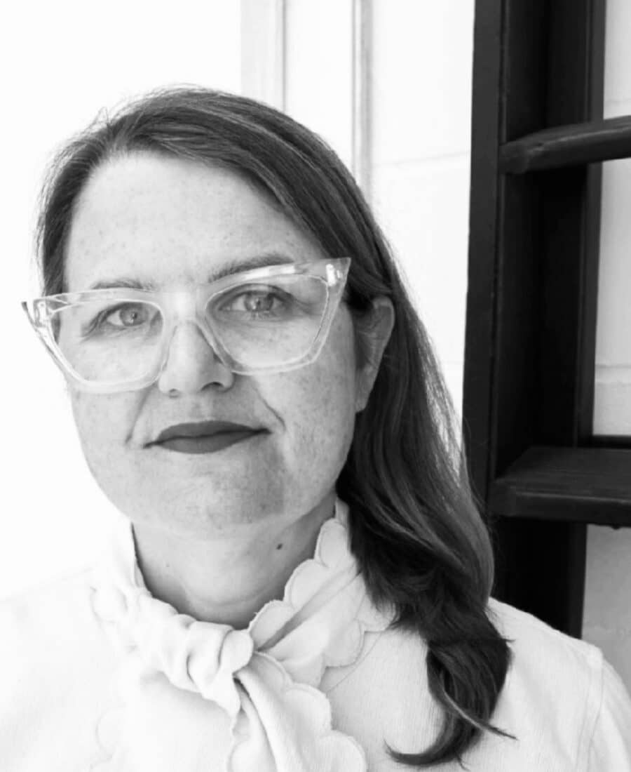 Woman with glasses and a ponytail, wearing a scallop-edged top, stands next to a dark wooden ladder. Black and white photo.