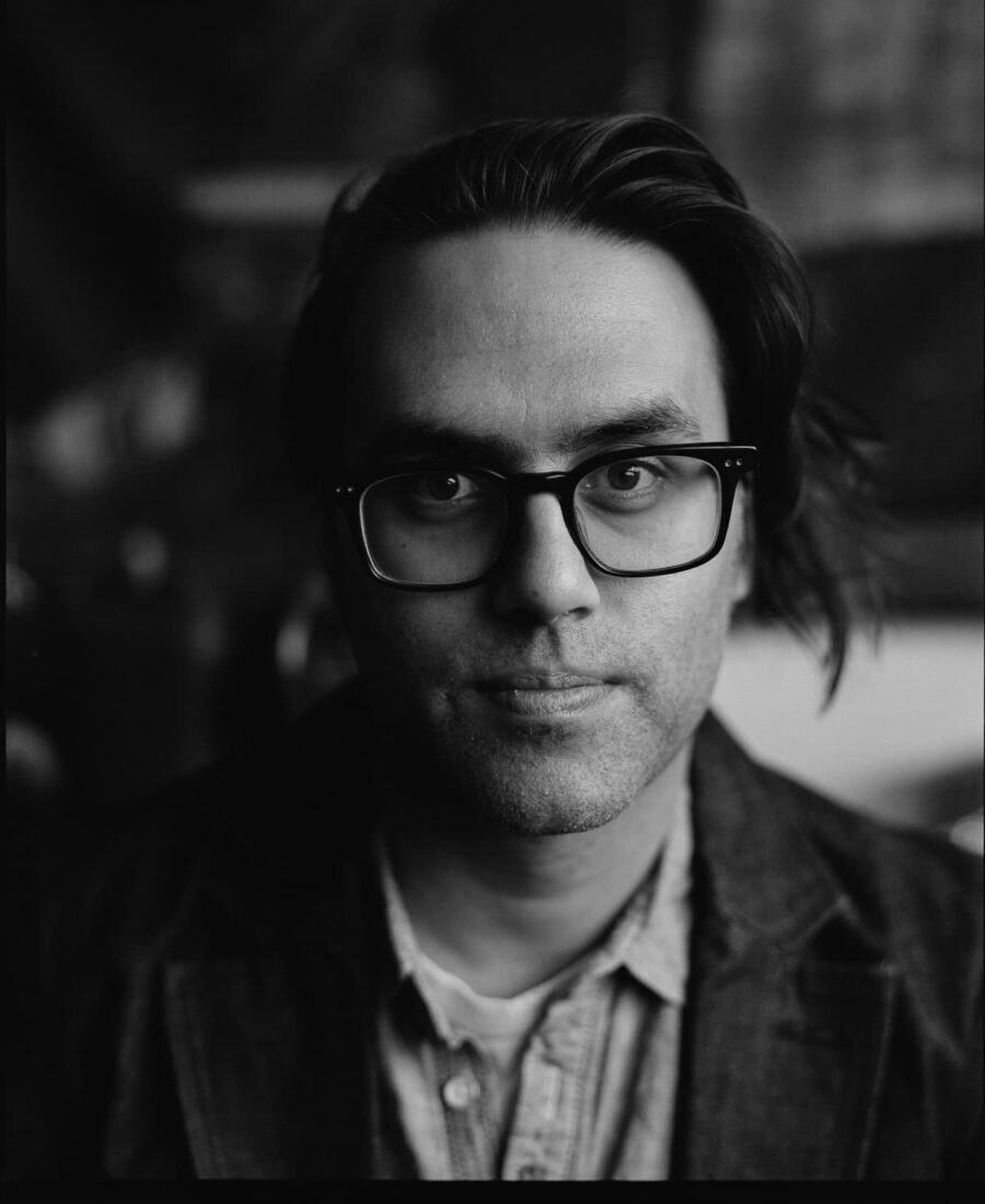 A person with glasses and long hair, wearing a denim shirt and blazer, looking directly at the camera in a black and white portrait.