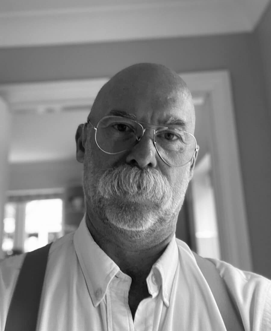 A person with glasses and a mustache is wearing a collared shirt and suspenders, standing indoors. The image is in black and white.