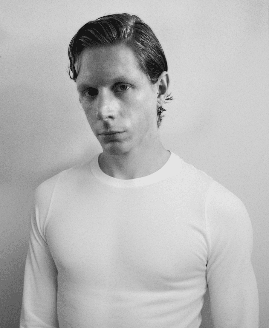 A person with short hair wearing a white long-sleeve shirt, standing against a plain wall and looking directly at the camera. The image is in black and white.