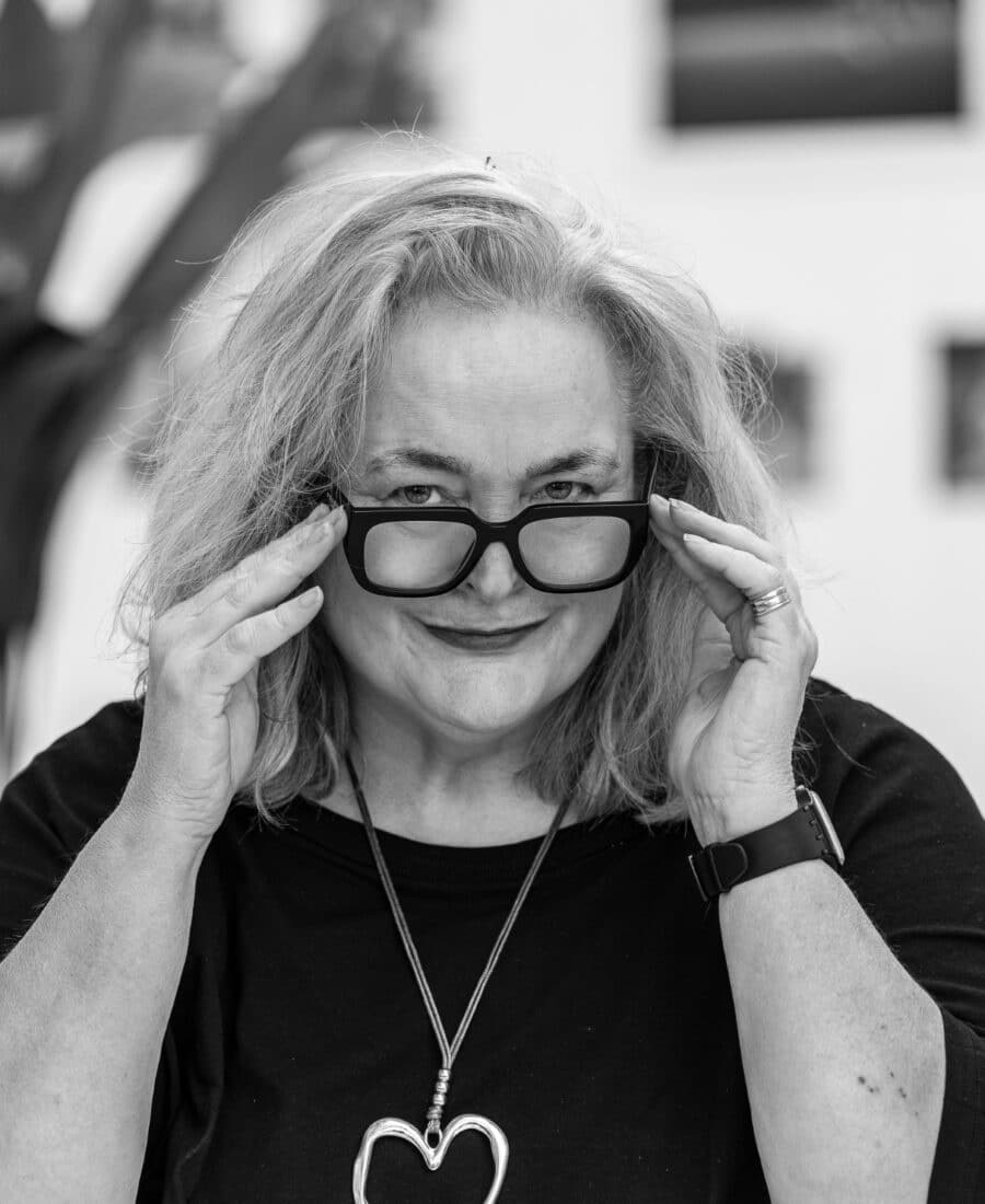 A person with shoulder-length hair and glasses smiles while adjusting their frames. They wear a dark top and a heart-shaped pendant. Black and white photo.