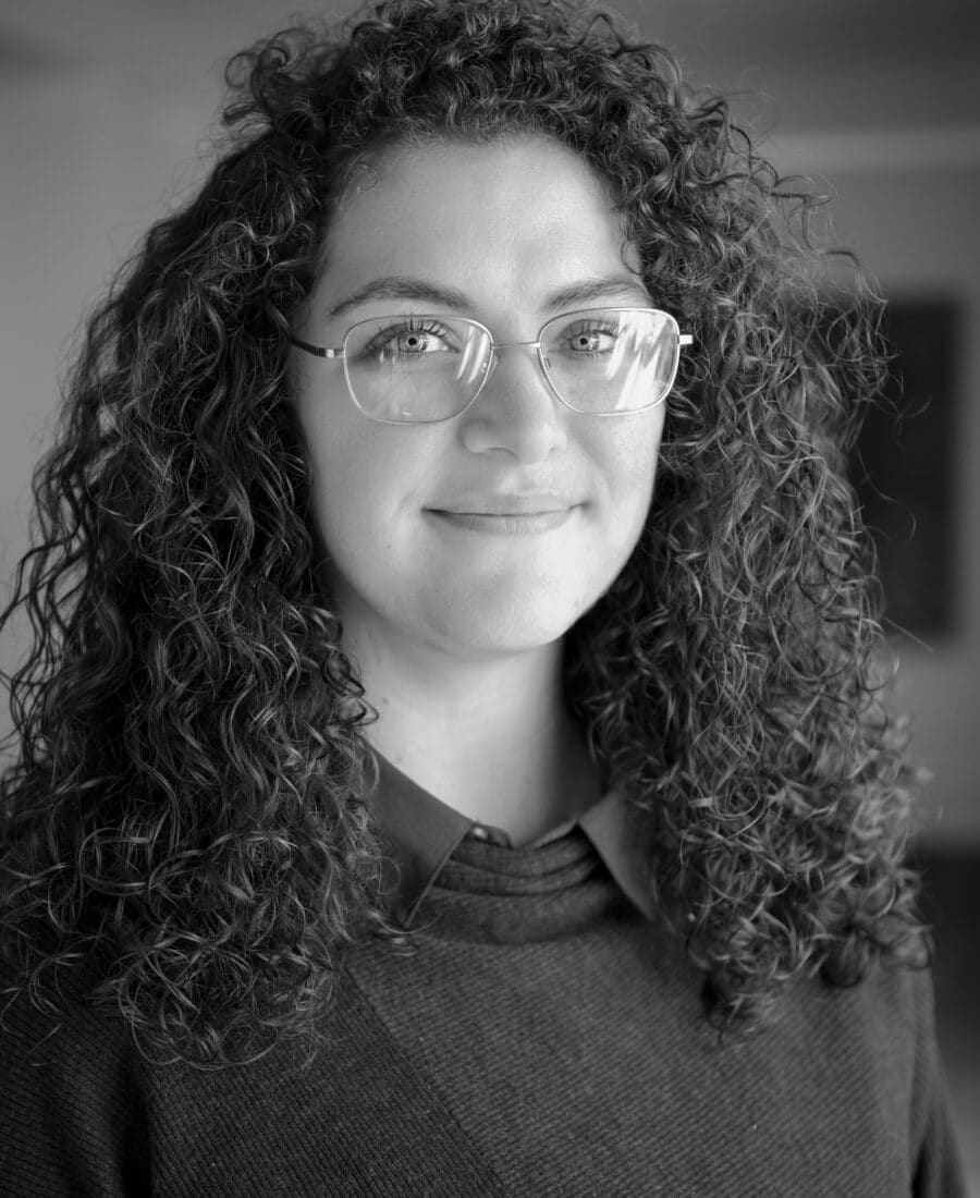 A person with curly hair and glasses is smiling while wearing a dark sweater. The image is in black and white.