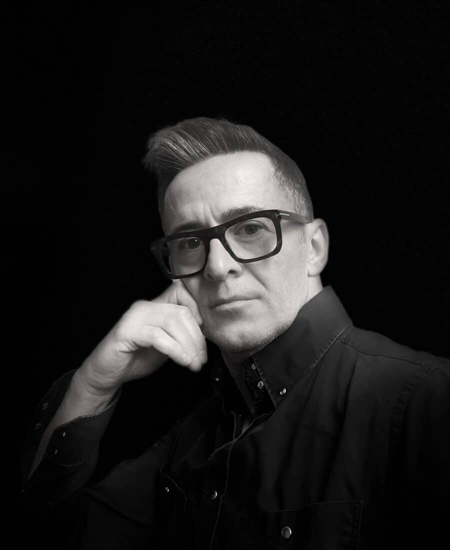 Man with short hair and glasses in black shirt, sitting against a dark background, with hand to chin.