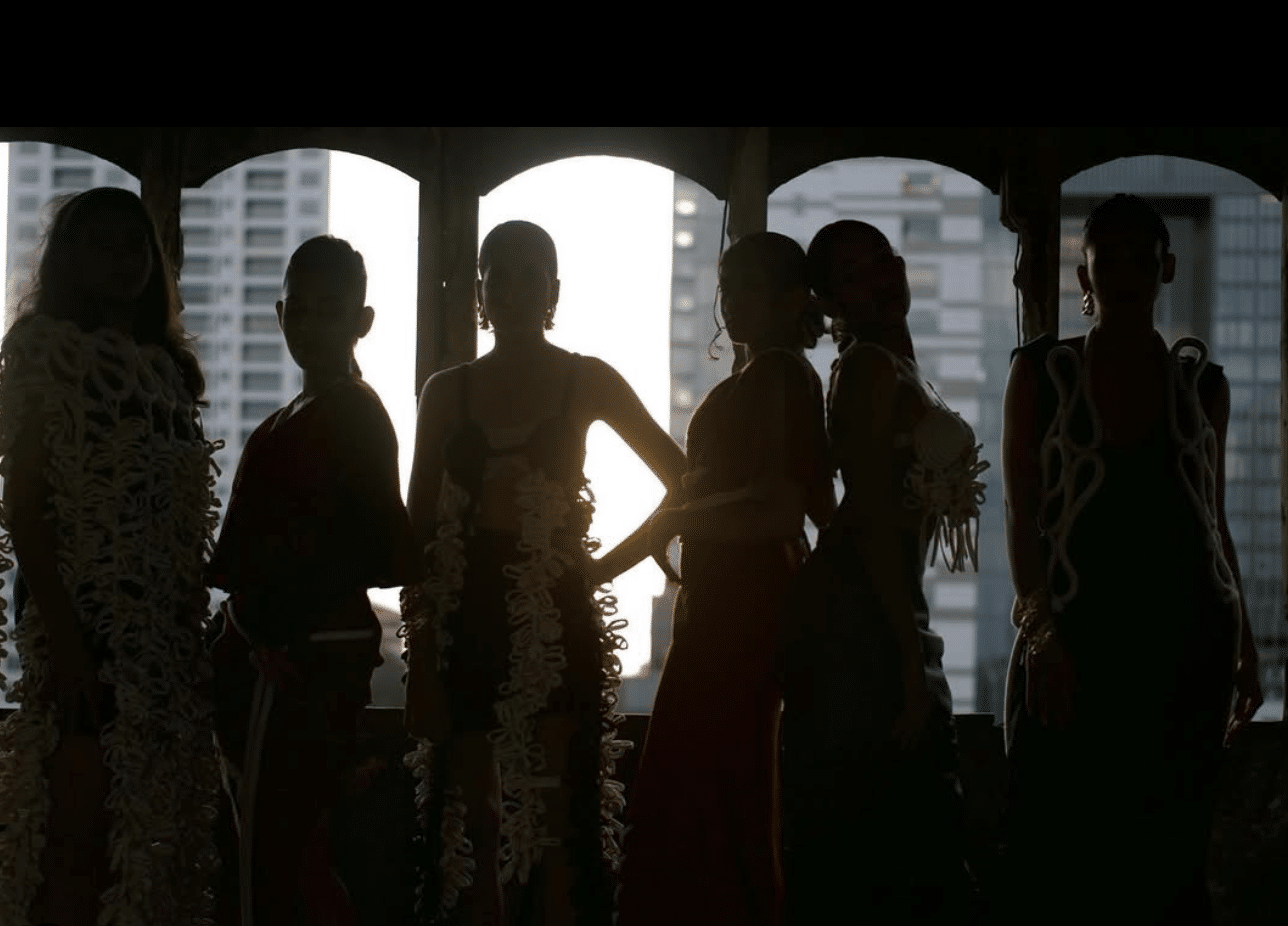 Silhouettes of six people standing in a line against a backlit urban skyline, wearing ornate outfits.